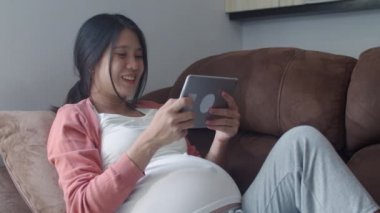 Hamilelik bilgileri için tablet kullanan Asyalı hamile bir kadın. Anne kendini mutlu hissederken pozitif ve huzurlu gülümserken çocuğuyla ilgilenirken oturma odasında kanepede uzanmış ev konseptinde..