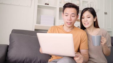 Dizüstü bilgisayar kullanarak ve sıcak fincan kahve oturma odasında evde içme Asya çift, tatlı çift kanepede yatarken aşk keyfini ne zaman evde dinlenin. Yaşam tarzı çift evde kavramı relax.