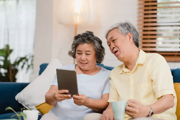 Asian elderly couple using tablet and drinking coffee in living room at home, couple enjoy love moment while lying on sofa when relaxed at home. Enjoying time lifestyle senior family at home concept. — Stock Photo, Image