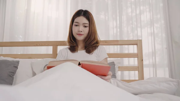 Vakker, ung asiatisk kvinne som leser en bok mens hun ligger på senga og slapper av på soverommet sitt om morgenen. Livsstilskvinner som bruker avslapningstid hjemme-konseptet . – stockfoto