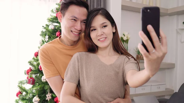 Couple asiatique en utilisant smartphone selfie avec arbre de Noël décorer leur salon à la maison au Festival de Noël. Lifestyle femme et homme heureux célébrer Noël d'été et concept de nouvelle année . — Photo