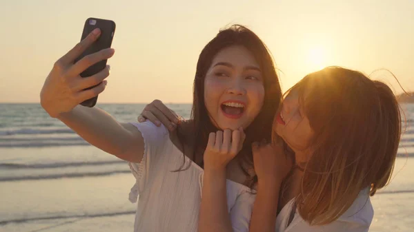 Młoda Azjatycka Para lesbijek za pomocą smartfona robienia selfie w pobliżu plaży. Piękne kobiety LGBT para szczęśliwy relaks cieszyć się chwila miłości, gdy zachód słońca wieczorem. Styl życia lesbijek para podróży na plaży koncepcji — Zdjęcie stockowe