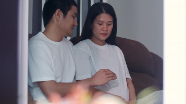 若いアジアの妊娠中のカップルの男性は 彼の子供と話して彼の妻の腹に触れます お母さんとお父さんは 赤ちゃんの世話をしながら 幸せな笑顔を感じ 自宅のリビングルームでソファに横たわって妊娠 スローモーション — ストック動画