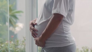 Genç Asya Hamile çift sarılmak ve çocukları ile konuşurken göbek tutarak. Anne ve baba mutlu gülümseyerek huzurlu bir şekilde bakarken bebek, evde oturma odasında pencerenin yanında hamilelik. Yavaş çekim.
