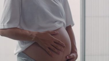 Genç Asya Hamile çift adam eşi göbek çocuğu ile konuşurken öpmek. Anne ve baba mutlu gülümseyerek huzurlu bir şekilde bakarken bebek, evde oturma odasında pencerenin yanında hamilelik. Yavaş çekim.