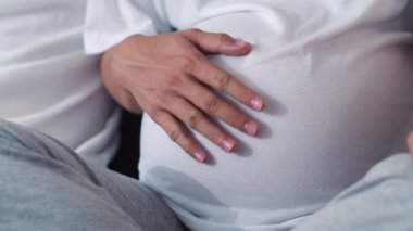 Genç Asya Hamile çift adam eşi karın çocuğu ile konuşurken dokunmak. Anne ve baba mutlu bir gülümseme huzurlu iken bakım bebek, hamilelik evde oturma odasında kanepeüzerinde yatan. Yavaş çekim.