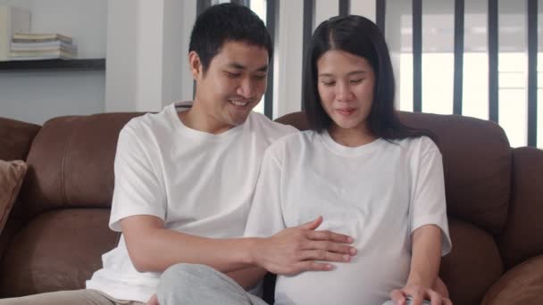 若いアジアの妊娠中のカップルは 腹を保持するハートサインを作る お母さんとお父さんは 世話をしながら穏やかな笑顔を感じ 自宅のコンセプトでリビングルームのソファに横たわって妊娠 スローモーション — ストック動画