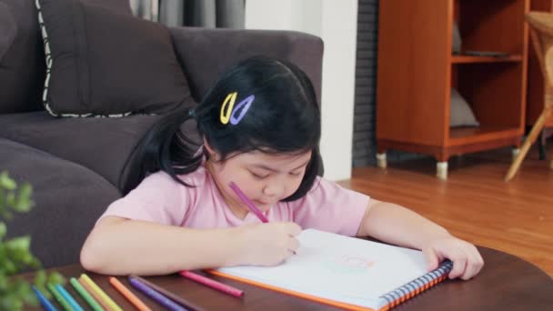 自宅で描く若いアジアの女の子 アジア日本の女性の子供は ソファに横たわってスケッチブックで楽しい幸せな描き漫画をリラックスして 朝のコンセプトでリビングルームで快適さと落ち着きを感じます スローモーション — ストック動画
