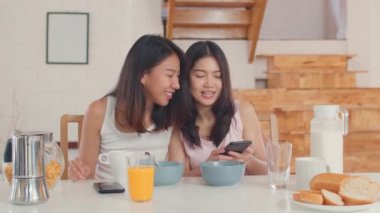 Asya lezbiyen LGBTQ kadınlar çift evde kahvaltı var, Genç Asya sevgilisi kızlar mutlu cep telefonu kontrol haberleri kullanarak İçecek suyu, Mısır gevreği ve mutfakta süt sabah kavramı.