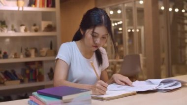 Asyalı öğrenci kadınlar üniversitede kütüphanede kitap okuyor. Genç lisans kız ev ödevi yapmak, ders kitabı okumak, üniversite kampüsünde ders masasında bilgi için çok çalışmak fazla mesai gece. Yavaş çekim.