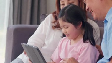 Asyalı dedesi ve torunu evde tablet kullanarak. Üst düzey Çinli, büyükbaba ve büyükanne mutlu genç kız ile aile dinlenmek geçirmek sosyal medya kontrol, oturma odasında kanepeüzerinde yatıyordu. Yavaş çekim.