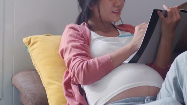 ノートに赤ちゃんを描く若いアジアの妊婦 お母さんは 自宅のコンセプトでリビングルームでソファに横たわっているケアの子供を取りながら 肯定的で平和な笑顔を幸せに感じています スローモーション — ストック動画