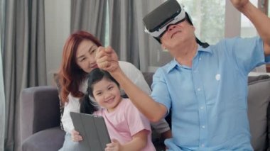 Asyalı dedesi ve torunu sanal gerçeklik ve tablet kullanarak evde oyun oynamak. Üst düzey Çinli, büyükbaba ve büyükanne oturma odasında kanepeye yatan genç kız ile mutlu dinlenmek. Yavaş çekim.