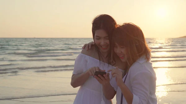 Jeune couple lesbienne asiatique utilisant un téléphone intelligent près de la plage. Belles femmes couple lgbt heureux se détendre profiter moment d'amour lorsque le coucher du soleil dans la soirée. Mode de vie lesbienne couple Voyage sur la plage concept — Photo