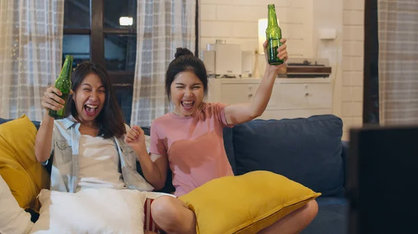 Lesbian lgbt women couple party at home, Asian female drinking beer watching TV cheer soccer funny moment together on sofa in living room in night. Young lover football fan, celebrate holiday concept.
