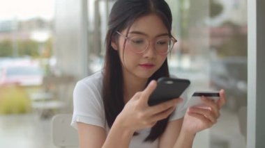 Serbest Asya kadın kahve dükkanında online alışveriş. Genç Asya Kız cep telefonu kullanarak, kredi kartı satın almak ve akşam konsepti açık kafede masada e-ticaret internet satın. Yavaş çekim.