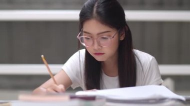 Asyalı öğrenci kadınlar üniversitede kütüphanede kitap okuyor. Üniversite kampüs konseptinde ders masasında bilgi için çok çalışırken genç lisans kız stres yorgun sorun var. Yavaş çekim.