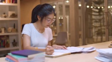 Asyalı öğrenci kadınlar üniversitede kütüphanede kitap okuyor. Genç lisans kız ev ödevi yapmak, ders kitabı okumak, üniversite kampüsünde ders masasında bilgi için çok çalışmak fazla mesai gece. Yavaş çekim.