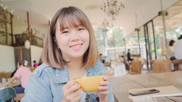 Frilans asiatisk kvinne som drikker varm kopp grønn te eller kaffe mens hun sitter på bordet i kafeen. Livsstilssmarte vakre kvinner slapper av i kaffebutikkkonsepter . – stockfoto