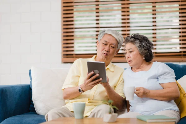 Asian elderly couple using tablet and drinking coffee in living room at home, couple enjoy love moment while lying on sofa when relaxed at home. Enjoying time lifestyle senior family at home concept. — Stock Photo, Image
