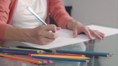 Genç Asyalı hamile kadın karnına ve ailesine not defterine bebek çiziyor. Anne kendini mutlu hissederken, pozitif ve huzurlu gülümserken çocuk evde oturma odasında kendine dikkat ederken..