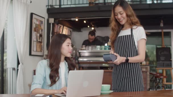 アジアの若いフリーランスの女性は コーヒーショップで非接触クレジットカードを支払います アジアの幸せな女の子バリスタウェイターは 顧客のためのクレジットカードリーダーマシンを保持する灰色のアポンを着用し カフェで技術で支払うことができます — ストック動画