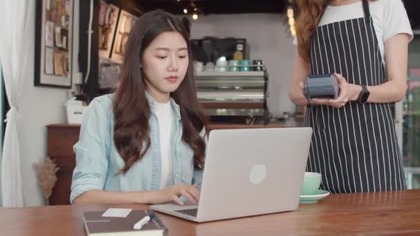 若いアジア人のフリーランス女性はコーヒーショップで非接触を支払う アジアの幸せな女性バリスタウェイターは カフェで携帯電話のスキャン支払いを使用して顧客のためのクレジットカードリーダーマシンを保持灰色のエプロンを着用 — ストック動画