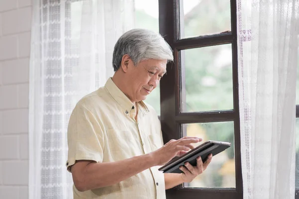 Asiatiska äldre man använder Tablet PC kontrollera sociala medier nära fönster i vardagsrum hemma. Livsstil Senior män hemma Concept. — Stockfoto