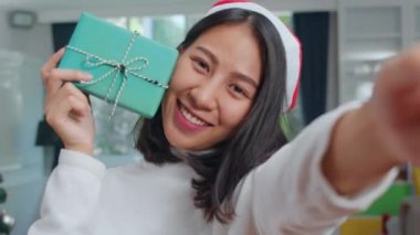 Asyalı kadınlar Noel festivalini kutluyor. Kadın genç Noel şapka giymek mutlu holding Hediye ve Noel ağacı ile akıllı telefon selfie kullanarak evde oturma odasında xmas kış tatilleri keyfini rahatlayın.