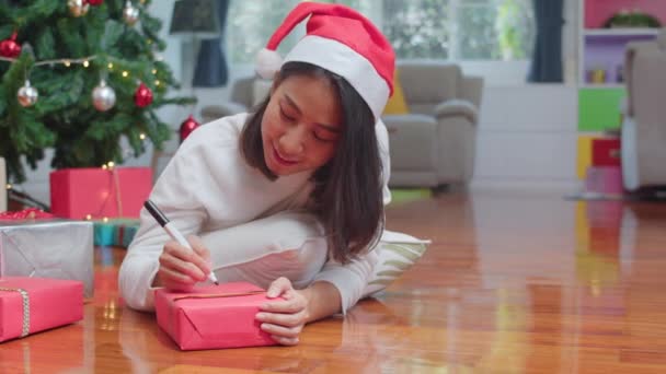 アジアの女性はクリスマスフェスティバルを祝います 女性の十代の若者はセーターとサンタ帽子を着用幸せにクリスマスツリーの近くの贈り物に願いを書いて幸せに書き込み 自宅のリビングルームで一緒にクリスマス冬の休日を楽しみます — ストック動画
