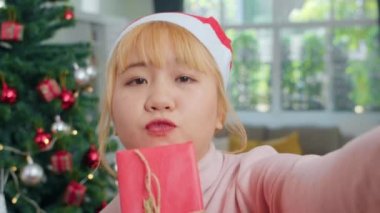 Asyalı kadınlar Noel festivalini kutluyor. Kadın genç Noel şapka giymek mutlu holding Hediye ve Noel ağacı ile akıllı telefon selfie kullanarak evde oturma odasında xmas kış tatilleri keyfini rahatlayın.