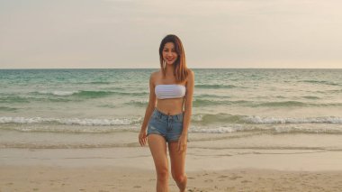Kum sahilinde yürüyen Asyalı bir kadın. Bikini genç mutlu kadın dinlenmek ve eğlenceli tropik deniz yakınında yürürken gün batımı sırasında tatil, tatil, yaz gezisi kavramı.