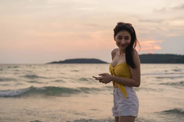 Азіатські жінки-підлітка ходьба на пляжі. Красива дівчина за допомогою мобільного телефону перевірки соціальних медіа біля моря, насолоджуючись її літнього відпочинку відпустку, коли захід сонця у вечірній час. — стокове фото