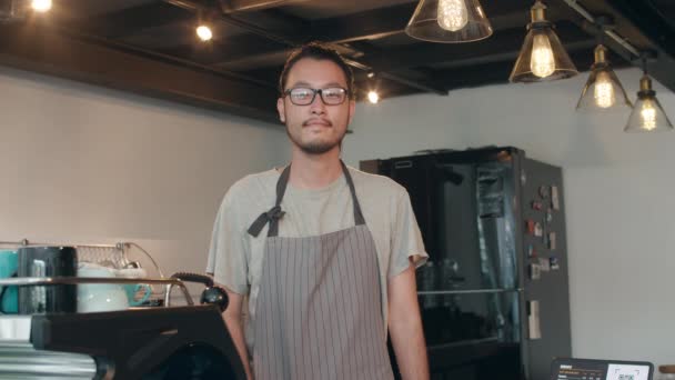 在城市咖啡店里 年轻的亚洲男子巴里斯塔面带微笑 面带微笑 小业主日本男人穿着围裙放松下来 微笑着看着站在咖啡店柜台前的相机 — 图库视频影像