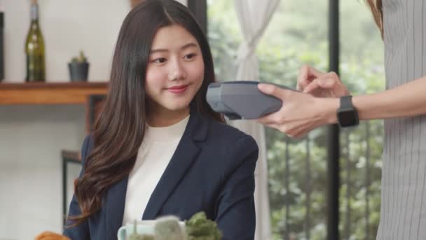 若いアジアのビジネス女性はコーヒーショップで非接触を支払う アジアの幸せな女の子バリスタウェイターは カフェで携帯電話のスキャン支払いを使用して顧客のためのクレジットカードリーダーマシンを保持グレーエプロンを着用 — ストック動画