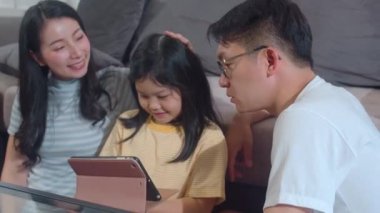 Genç Asyalı aile ve kızı evde tablet kullanmaktan mutlu. Japon anne ve baba, oturma odasında kanepede uzanmış film izleyen küçük kızla rahatlar. Komik ebeveyn ve sevimli çocuk eğleniyor..