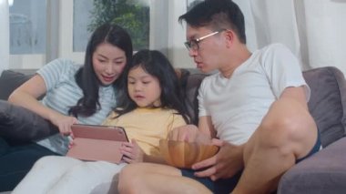 Genç Asyalı aile ve kızı evde tablet kullanmaktan mutlu. Japon anne ve baba, oturma odasında kanepede uzanmış film izleyen küçük kızla rahatlar. Komik ebeveyn ve sevimli çocuk eğleniyor..