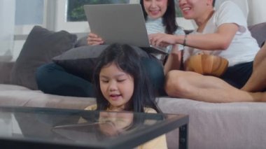 Genç Asyalı aile ve kız evde tablet ve dizüstü bilgisayar kullanmaktan mutlu. Japon anne ve baba, küçük kızın film izlemesi ve oturma odasındaki kanepede uzanan sosyal medyayı kontrol etmesi ile rahatlar..