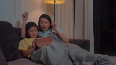 Genç Asyalı aile ve kızı evde tablet kullanmaktan mutlu. Koreli anne, geceleri evdeki oturma odasında uyumadan önce kanepede uzanmış film izleyen küçük kızla rahatlıyor..
