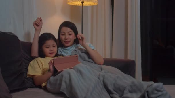 若いアジアの家族や娘は自宅でタブレットを使用して幸せ 韓国のお母さんリラックスとともに女の子見る映画あります上のソファ前に寝ますで家の中で夜 — ストック動画
