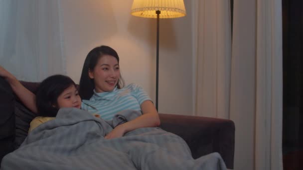 若いアジアの家族や娘は夜に自宅でテレビを見て 家族の時間を使って幸せな女の子と韓国の母親はリビングルームでソファに横にリラックスします 面白いお母さんと素敵な子供が楽しんでいます — ストック動画