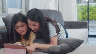 Genç Asyalı aile ve kızı evde tablet kullanmaktan mutlu. Japon anne, evdeki oturma odasındaki kanepede uzanmış film izleyen küçük kızla rahatlıyor. Komik anne ve güzel çocuk eğleniyor..