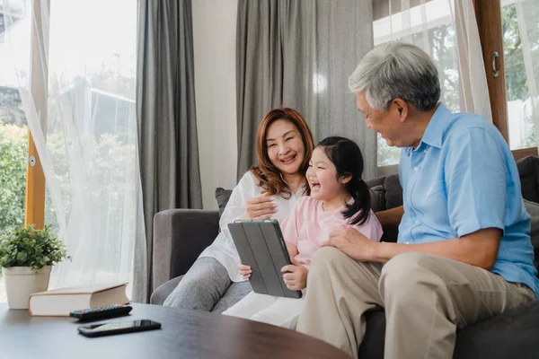 Asian grandparents and granddaughter using tablet at home. Senior Chinese, grandpa and grandma happy spend family time relax with young girl checking social media, lying on sofa in living room concept — Stock Photo, Image