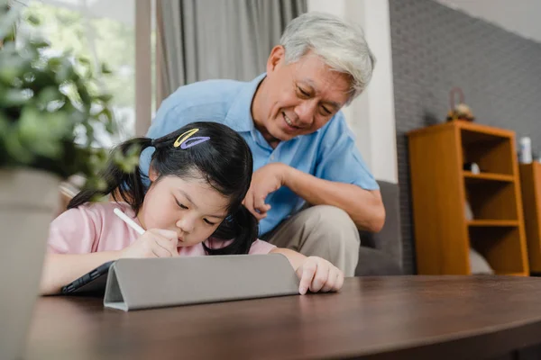 El abuelo asiático enseña a su nieta a dibujar y hacer deberes en casa. Senior chino, abuelo feliz relajarse con la joven acostada en el sofá en la sala de estar en el concepto de casa. — Foto de Stock