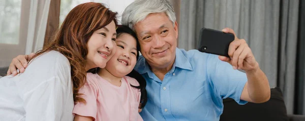 Asiático abuelos selfie con nieta en casa. Senior chino feliz pasar tiempo en familia relajarse con el teléfono móvil con el niño joven acostado en el sofá en la sala de estar. Fondo de la bandera panorámica . — Foto de Stock