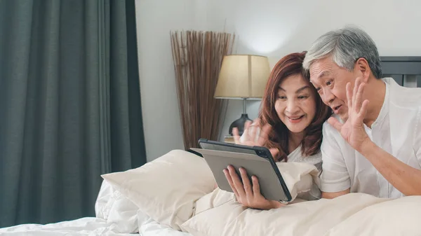 Asian senior couple using tablet at home. Asian Senior Chinese grandparents, video call talking with family grandchild kids while lying on bed in bedroom at home in the morning concept. — Stock Photo, Image