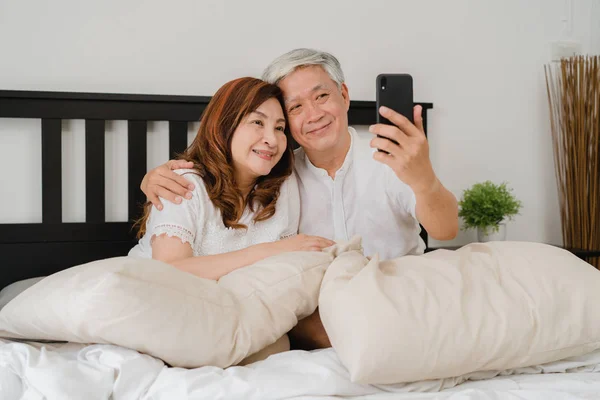Asiática pareja mayor selfie en casa. Asiático Senior abuelos chinos, marido y esposa feliz usando el teléfono móvil selfie después de despertar acostado en la cama en el dormitorio en casa en el concepto de la mañana. — Foto de Stock