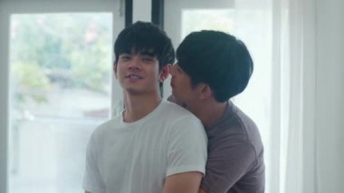 Asyalı eşcinsel çift evlerinin penceresinin önünde durup sarılıyorlar. Genç Asyalı Lgbtq + erkekler mutlu öpüşürler birlikte dinlenirler modern evdeki oturma odasında romantik zaman geçirirler..