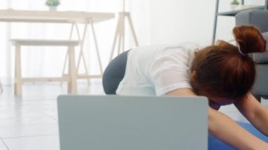 Spor giyinen genç Koreli bayan evde yoga videosu izlemek için spor yapıyor ve dizüstü bilgisayar kullanıyor. Kişisel antrenörle uzak eğitim, sosyal mesafe, çevrimiçi eğitim kavramı.