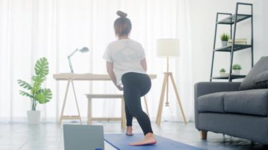 Spor giyinen genç Koreli bayan evde yoga videosu izlemek için spor yapıyor ve dizüstü bilgisayar kullanıyor. Kişisel antrenörle uzak eğitim, sosyal mesafe, çevrimiçi eğitim kavramı.